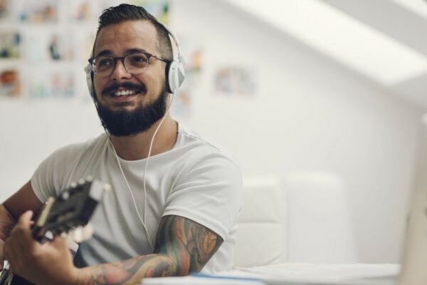 A bearded man smiles while wearing headphones and glasses, he is playing an acoustic guitar