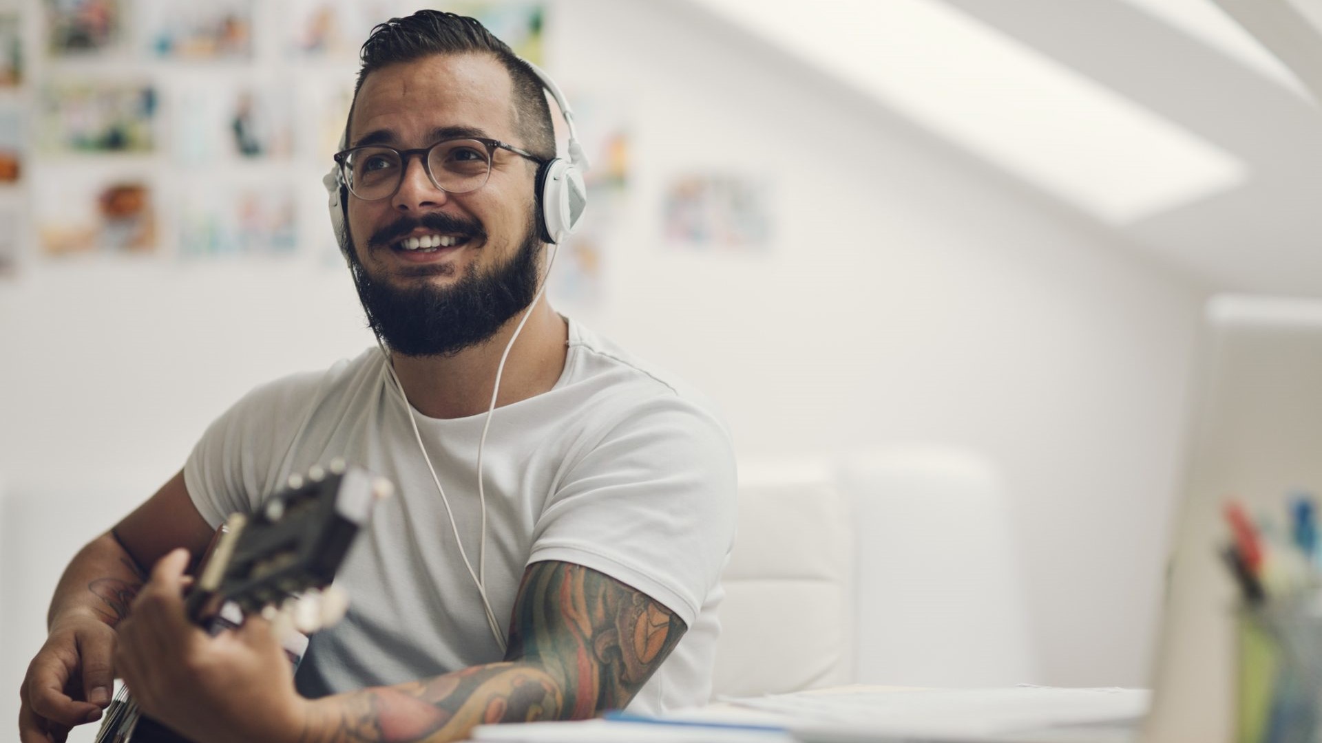 A bearded man smiles while wearing headphones and glasses, he is playing an acoustic guitar