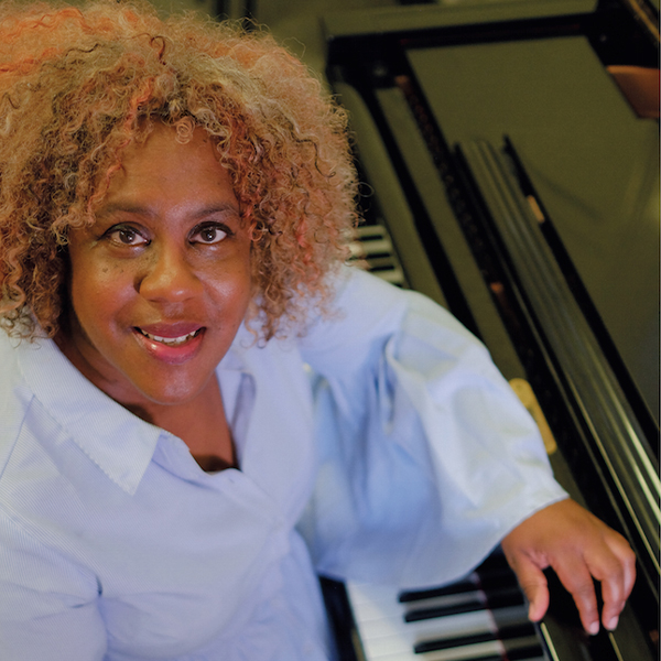 Errollyn Wallen, a black woman with thick curly hair with streaks of blonde, red and brown, she smiles up at the camera from her seated position at a piano