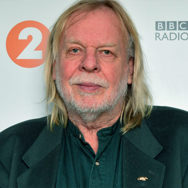 Rick Wakeman, an older white man with a grey beard and moustache and shoulder length thin blonde hair, he is wearing a dark green