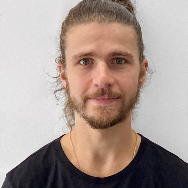 Sebastian Willis, a young man, with a light brown wispy beard and long hair tied up in a bun on the back of his head. He is looking directly into the camera and wearing a black t shirt.