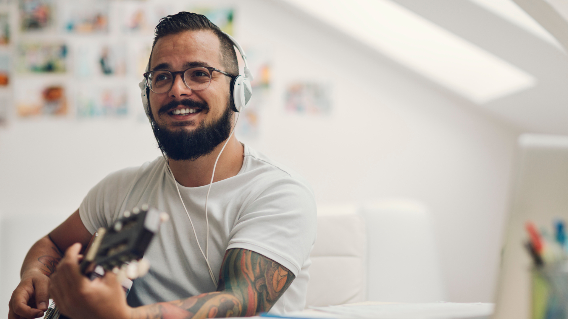 A man with a large beard and moustache and gelled hair looks away from the camera, he is playing an acoustic guitar and wearing white headphones.