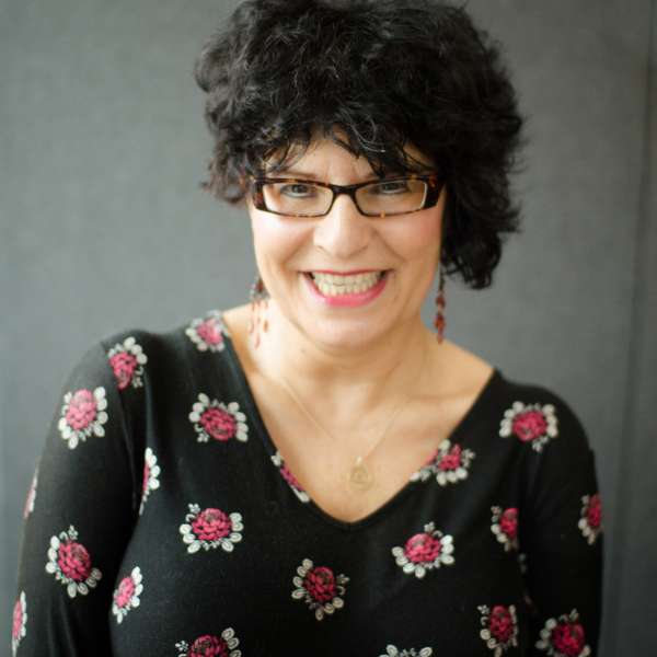 Athena Pite, a smiling lady wearing bright pink lipstick, tortoise shell glasses, her dark black hair is short but thick and full. She is wearing a black top with pink flowers on.