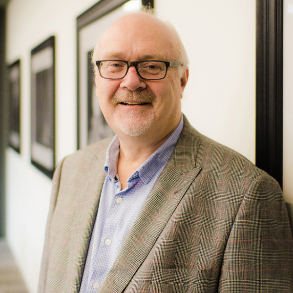 John Logan, an older man with short grey stubble wearing black glasses, a tweed jacket and blue shirt