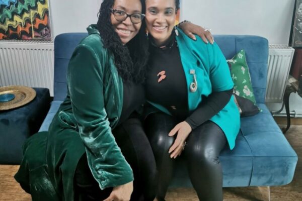 Two women with black hair, both wearing green jackets sat on a small sofa, the lady on the left is wearing glasses and has her arm around the other, they are both smiling and happy.