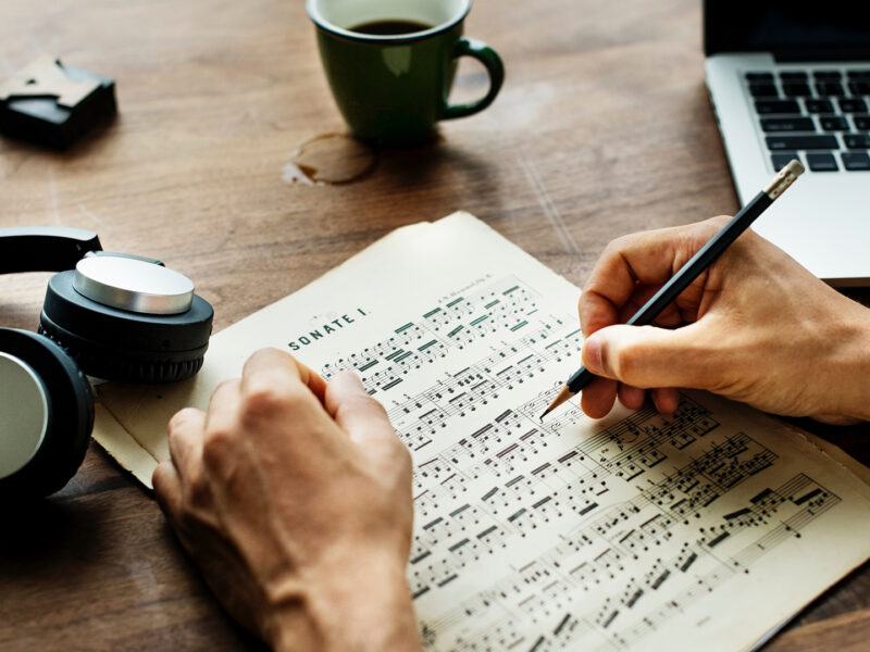 A hand writing on sheet music with pencil, a laptop, headphones and coffee cup are on the table