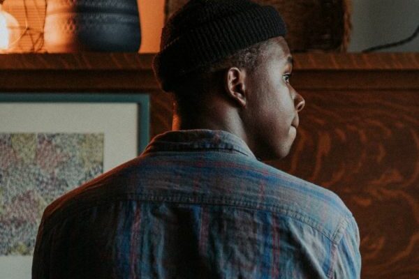 A young black man looking out to the right as he sits in front of a brown piano, he is waring a muted blue check shirt and black wool hat.