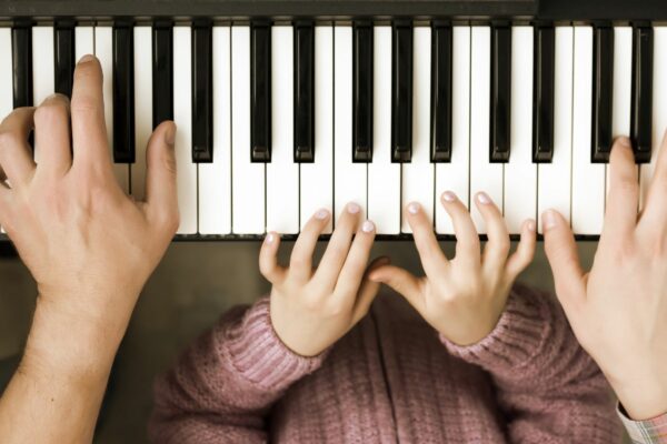 Two sets of hands playing a piano, one is a small child who is wearing a pink jumper and silver nail polish, one is an adult wearing a checked shirt.