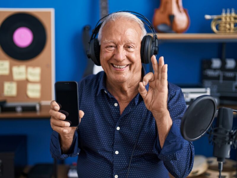 An older man with grey hair wearing headphones, smiling and holding up a phone in one hand and the OK signal with the other, he is sat in a recording studio